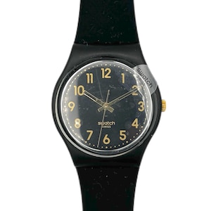 May include: A black Swatch watch with a black dial and gold-colored hands and numerals. The watch has a black strap and a gold-colored crown. The watch face is protected by a clear plastic cover. The word "Swatch" is printed on the watch face.