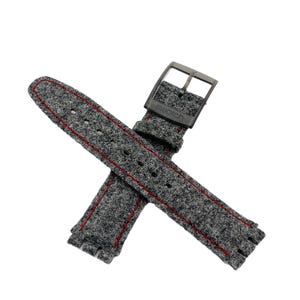 Puede incluir: Una correa de reloj gris con costuras rojas. La correa está hecha de un material texturizado y presenta una hebilla de metal con el nombre de la marca "Swatch" grabado. La correa está diseñada para sujetarse a un reloj.