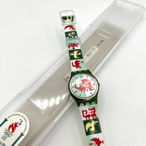 May include: A Swatch quartz watch with a green strap featuring colorful animal and geometric designs. The watch face displays an elephant graphic. The watch is in its original clear plastic case, with the Centennial Olympic Games logo visible.