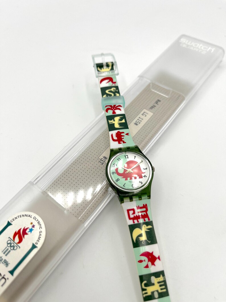 May include: A Swatch quartz watch with a green strap featuring colorful animal and geometric designs. The watch face displays an elephant graphic. The watch is in its original clear plastic case, with the Centennial Olympic Games logo visible.