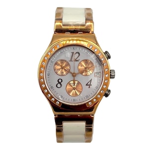 May include: A gold-toned Swatch Swiss watch with a white face, featuring gold subdials and a date display. The bezel is adorned with small crystals. The watch has a white and gold band.