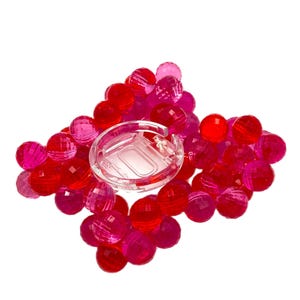 Puede incluir: Un pendiente de plástico transparente con cierre de bisagra, rodeado de un grupo de cuentas facetadas y translúcidas en tonos rojos y rosas. Las cuentas están dispuestas en forma de diamante, creando una exhibición visualmente atractiva.
