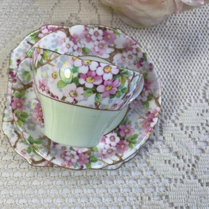 May include: A vintage teacup and saucer set with a floral pattern. The cup and saucer are both green with a white background and feature pink and white flowers. The saucer has a scalloped edge.