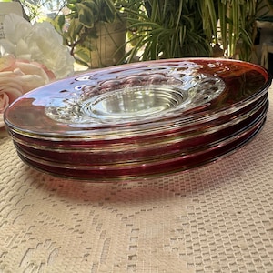 May include: Four red and clear glass plates are stacked. Each plate has a wide, red-tinted rim and a clear, circular centre. The plates are stacked on a white lace tablecloth, with a blurred background of flowers and greenery.