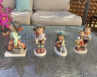 Op de afbeelding: Vier Hummel-beeldjes worden op een glazen tafel getoond. De beeldjes beelden kinderen uit in verschillende poses, waaronder een met een lam, een ander met een eend en twee kinderen die een mand vasthouden. De beeldjes zijn geschilderd in gedempte kleuren.