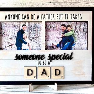 May include: A white wooden picture frame with a black border and two photo openings. The frame has the text "Anyone can be a father but it takes someone special to be a dad" and the word "DAD" spelled out with wooden letter tiles. The frame is standing on a white surface.