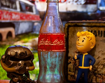 May include: A glass bottle of Nuka-Cola with a red and white label. The bottle is sitting on a wooden table with a Fallout Vault Boy figurine and bottle caps.