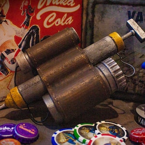 May include: A brown and silver Fallout Pip-Boy weapon replica with a yellow tip and a black leather strap. The weapon is lying on a table with a Vault-Tec logo and a figure of a Vault Boy giving a thumbs up.