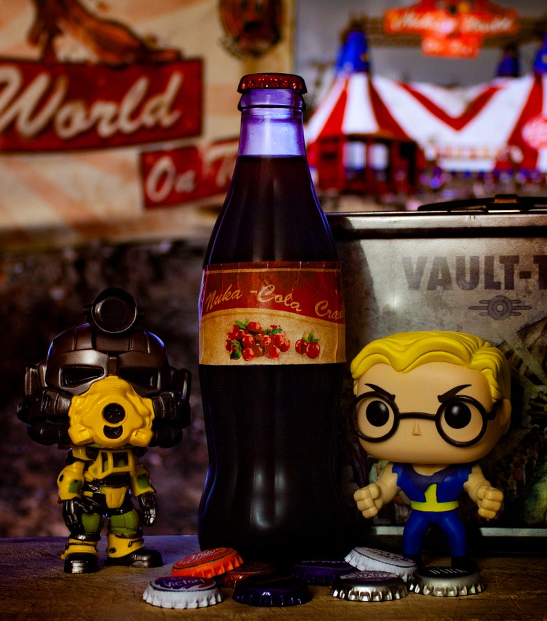 K&ouml;nnte beinhalten: Eine Glasflasche Nuka-Cola Cranberry Soda mit einem roten und wei&szlig;en Etikett. Die Flasche steht neben einer kleinen, gelben und schwarzen Roboterfigur und einer kleinen, gelben und blauen Figur mit Brille. Im Hintergrund befindet sich ein Schild mit der Aufschrift "World of Coca-Cola" und ein rot-wei&szlig; gestreiftes Zelt.