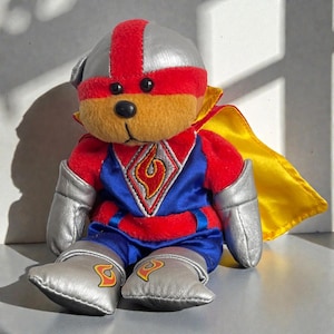 May include: A teddy bear toy dressed as a superhero. The bear has a silver helmet with a red stripe, a red and blue uniform with a flame emblem, silver boots and gloves, and a yellow cape. The bear is sitting on a white surface.