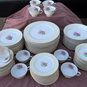 May include: A set of white china dinnerware with a delicate pink rose design and gold trim. The set includes dinner plates, salad plates, bowls, and teacups.