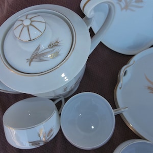 Peut inclure: Une théière en porcelaine blanche avec une bordure dorée et un motif de blé. La théière a un couvercle avec un bouton au centre. Il y a aussi quatre tasses à thé en porcelaine blanche avec une bordure dorée et un motif de blé.