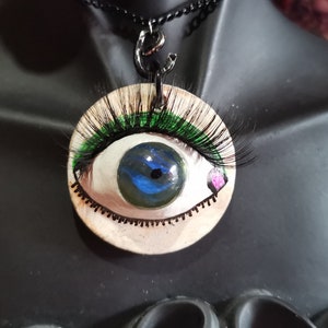 May include: A necklace with a round pendant featuring a painted eye with green eyelashes and a blue iris. The pendant is on a black chain.