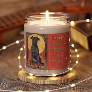 May include: A brown glass candle with a label that reads "Cane Corso" and "The Bouncer". The label features a black dog illustration and text describing the dog's personality. The candle is lit and sitting on a wooden coaster.