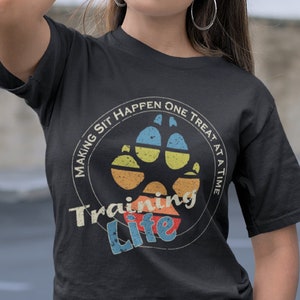 Pode incluir: Camiseta preta com um gráfico vintage de uma pegada de pata e o texto "Making Sit Happen One Treat At A Time Training Life".