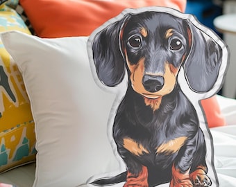 May include: A decorative pillow shaped like a dachshund dog. The pillow features a detailed illustration of the dog with black, brown, and tan fur. The dog is sitting upright with its ears flopped down. The pillow is on a white surface.