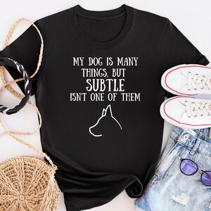 May include: Black t-shirt with white text that reads "My dog is many things, but subtle isn't one of them". A white silhouette of a dog's head is below the text.