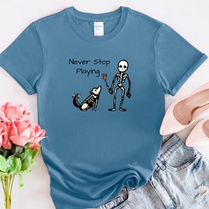 Może przedstawiać: Jasnoniebieska koszulka z nadrukiem szkieletu i psa grającego w frisbee. Tekst na koszulce brzmi "Never Stop Playing".
