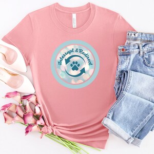 Könnte beinhalten: Hellrosa T-Shirt mit einem kreisförmigen Design, das "Interrupt & Redirect" mit einem Pfotenabdruck und Pfeilen zeigt. Das Design befindet sich in einem hellblauen Kreis mit Bokeh-Effekt. Das Shirt wird mit Jeans und weißen Schuhen kombiniert.