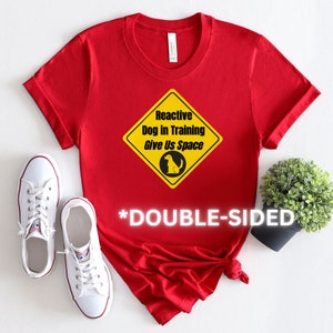 Op de afbeelding: Rood T-shirt met een geel ruitvormig bord met de tekst "Reactive Dog in Training Give Us Space" en een klein afbeelding van een hond. De tekst "*DOUBLE-SIDED*" is onder het bord gedrukt.