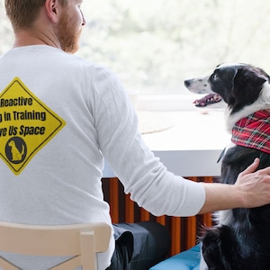 Op de afbeelding: Een man draagt een wit longsleeve shirt met een geel ruitvormig bord waarop staat "Reactive Dog in Training Give Us Space". Een zwart-witte hond met een rood-zwart geruit bandana zit naast hem.