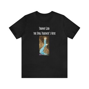Può includere: Una t-shirt nera a maniche corte con il testo bianco "THANK GOD THE DOG TRAINER'S HERE" sopra un grafico rettangolare che mostra una persona in un lungo abito blu inginocchiata con le braccia sollevate verso il cielo su sfondo scuro.