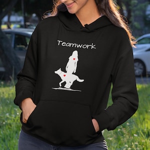 May include: Black hoodie with a white silhouette of a person and a dog walking side-by-side. The text "Teamwork" is printed in white above the silhouettes. Both the person and the dog have a red heart on their chest.