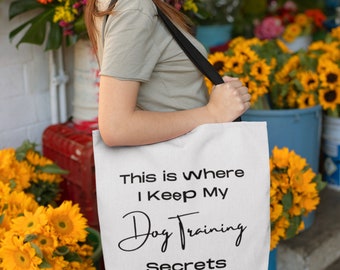 May include: White tote bag with black shoulder straps. The bag features the text "This is Where I Keep My Dog Training Secrets" in black. The background includes colorful flowers.
