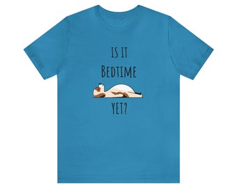 Op de afbeelding: Turquoise T-shirt met korte mouwen met de tekst "IS IT BEDTIME YET?" boven een cartoon illustratie van een liggende hond. De hond is bruin, wit en zwart.