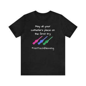 Op de afbeelding: Zwart T-shirt met de tekst "May all your catheters place on the first try" en de hashtag "#VetTechBlessing". Het shirt heeft drie kleurrijke katheter illustraties in roze, blauw en groen.