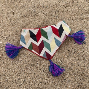 May include: A colorful dog bandana with a geometric pattern in red, black, green, blue, and beige. The bandana has a red border and is adorned with purple and blue tassels at each end. The item is displayed on a sandy surface.