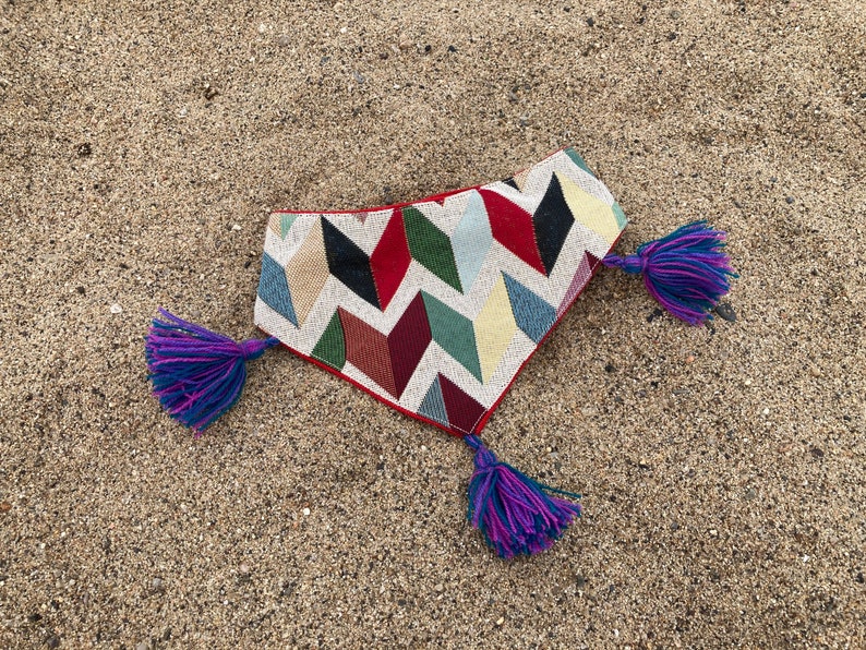 May include: A colorful dog bandana with a geometric pattern in red, black, green, blue, and beige. The bandana has a red border and is adorned with purple and blue tassels at each end. The item is displayed on a sandy surface.