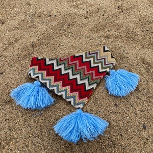 May include: A colorful, patterned clutch with a zig-zag design in red, white, and green, accented with three light blue tassels. The clutch is lying on a sandy surface.