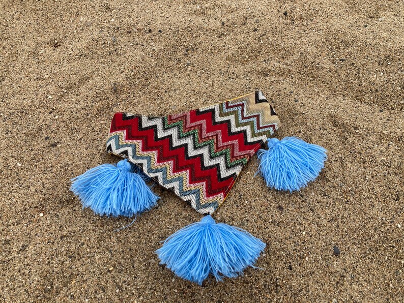May include: A colorful, patterned clutch with a zig-zag design in red, white, and green, accented with three light blue tassels. The clutch is lying on a sandy surface.