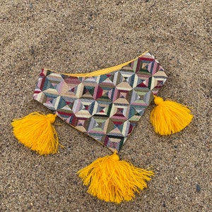 May include: A colorful geometric patterned pouch with a yellow zipper and three bright yellow tassels. The pouch is made of woven fabric and is resting on a sandy surface.