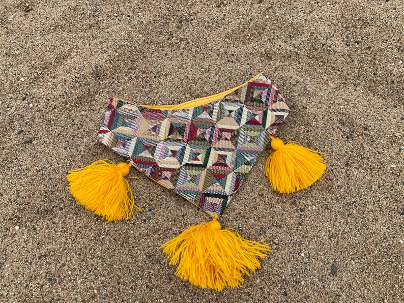 May include: A colorful geometric patterned pouch with a yellow zipper and three bright yellow tassels. The pouch is made of woven fabric and is resting on a sandy surface.