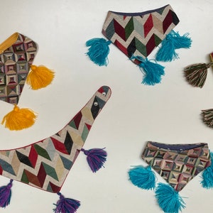 May include: A collection of patterned fabric bandanas with colorful tassels. The bandanas feature geometric and chevron designs in various colors, including yellow, blue, green, red, and purple. The tassels are attached to the corners of the bandanas.