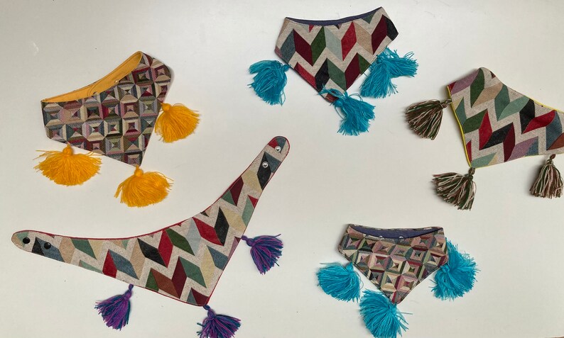 May include: A collection of patterned fabric bandanas with colorful tassels. The bandanas feature geometric and chevron designs in various colors, including yellow, blue, green, red, and purple. The tassels are attached to the corners of the bandanas.