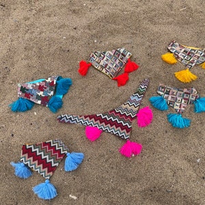 May include: An assortment of colorful, patterned fabric headbands with tassel accents. The headbands feature geometric and zigzag designs in various colors, including red, blue, yellow, and pink. They are arranged on a sandy surface.