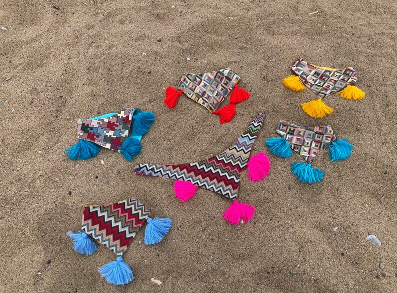 May include: An assortment of colorful, patterned fabric headbands with tassel accents. The headbands feature geometric and zigzag designs in various colors, including red, blue, yellow, and pink. They are arranged on a sandy surface.