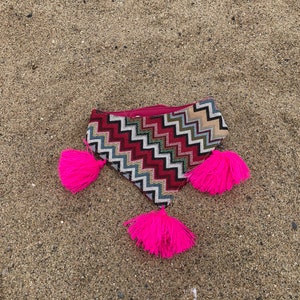 May include: A patterned clutch with a zigzag design in red, white, and blue, accented with bright pink tassels. The clutch is lying on a sandy surface, showcasing its vibrant colors and unique texture.