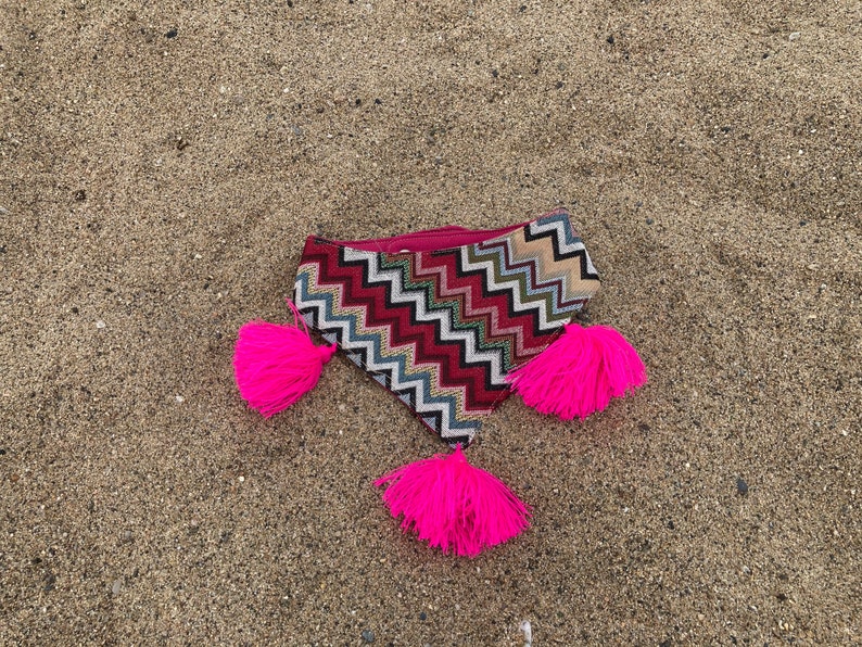 May include: A patterned clutch with a zigzag design in red, white, and blue, accented with bright pink tassels. The clutch is lying on a sandy surface, showcasing its vibrant colors and unique texture.
