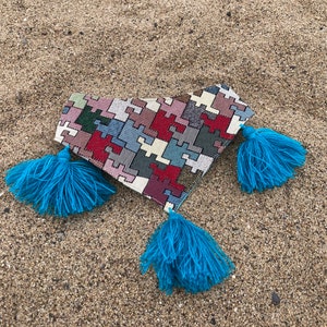 May include: A heart-shaped clutch with a puzzle-piece pattern in red, white, blue, and gray. The clutch has three bright blue tassels attached. The item is lying on a sandy surface.