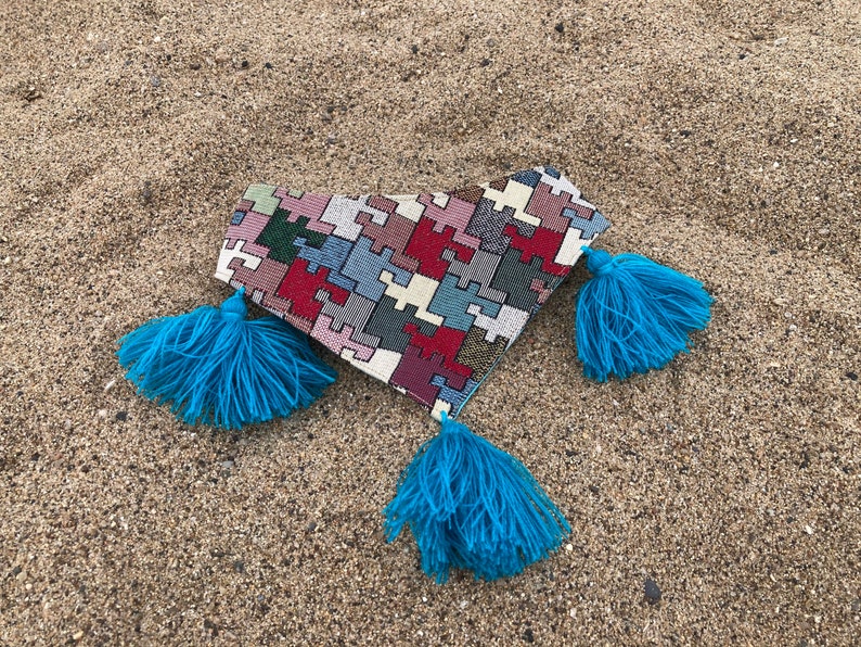 May include: A heart-shaped clutch with a puzzle-piece pattern in red, white, blue, and gray. The clutch has three bright blue tassels attached. The item is lying on a sandy surface.