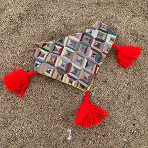 May include: A decorative textile with a geometric pattern in shades of green, blue, red, and beige, trimmed with yellow and adorned with three bright red tassels. The textile is lying on a sandy surface.