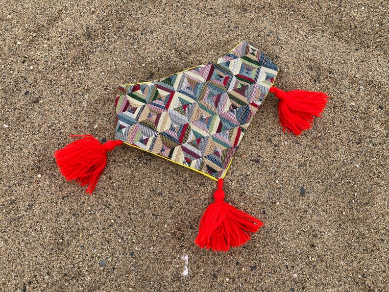 May include: A decorative textile with a geometric pattern in shades of green, blue, red, and beige, trimmed with yellow and adorned with three bright red tassels. The textile is lying on a sandy surface.