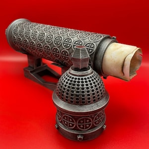 May include: A silver metal decorative box with a scroll inside. The box has a detailed design of circles and crosses. The box is on a red surface.