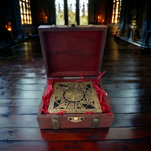 May include: An open wooden box with a brass latch, revealing a red and gold puzzle box with a circular design. The box is sitting on a wooden surface.