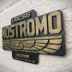 May include: A black and gold 3D printed sign with the text "USCSS NOSTROMO 180286 WETLAND-YUTANI CORPORATION"