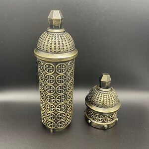 May include: Two gold-colored decorative containers with intricate patterns. The larger container is taller and has a more ornate design. The smaller container is shorter and has a simpler design.